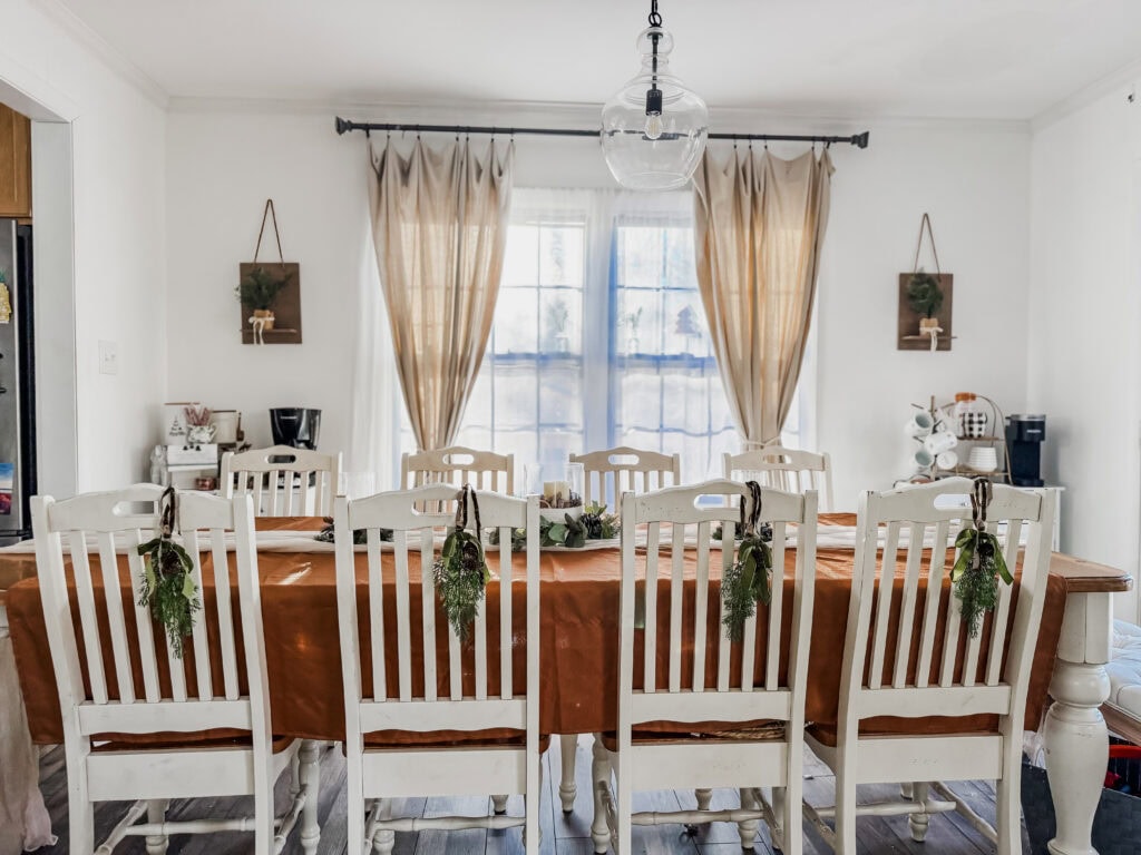 Festive dining room with a long table decorated with holiday greenery and a burnt orange tablecloth, surrounded by white chairs, with natural light from large windows and cozy curtains.