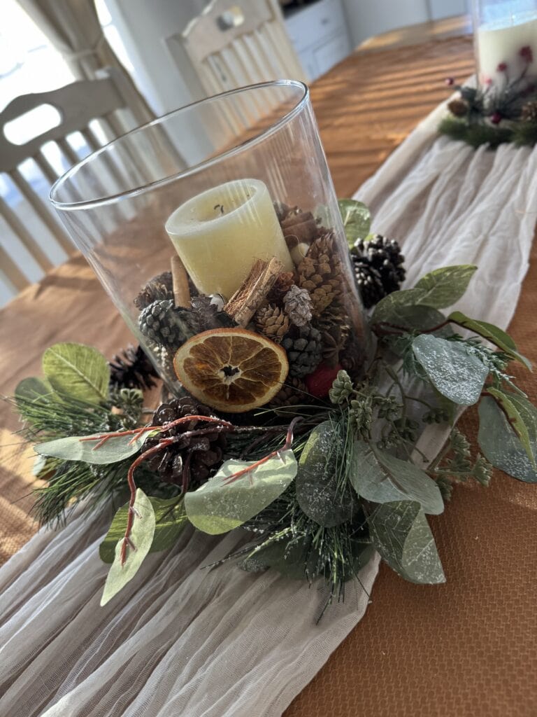 Vase centerpiece with candles, pinecones, dried orange slices, and greenery on a wooden table, perfect for holiday or winter decor.