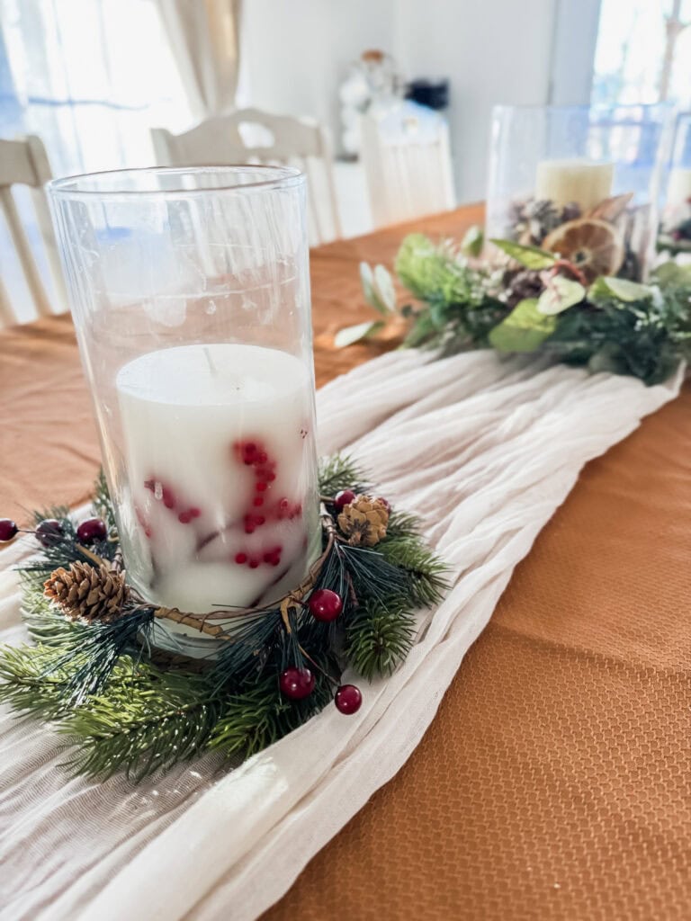 Festive holiday candle centerpiece with pinecones, greenery, and red berries on a rustic table runner for Christmas decorating.