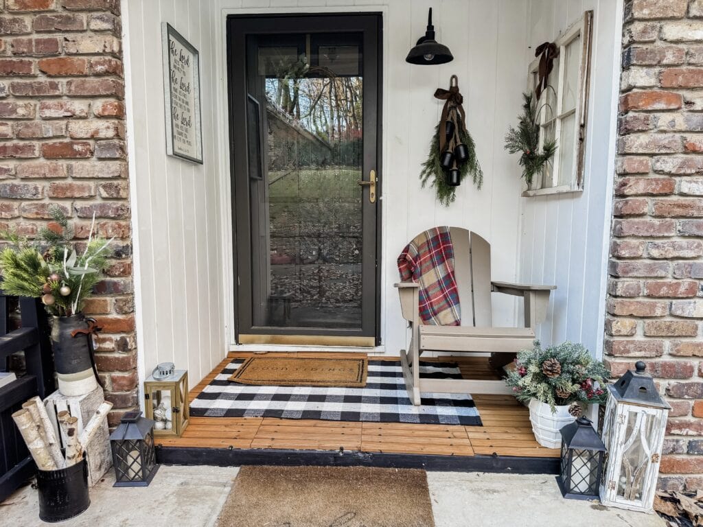 Cozy farmhouse porch decorated for Christmas with plaid blanket, greenery, lanterns, and rustic accents, creating welcoming holiday curb appeal.