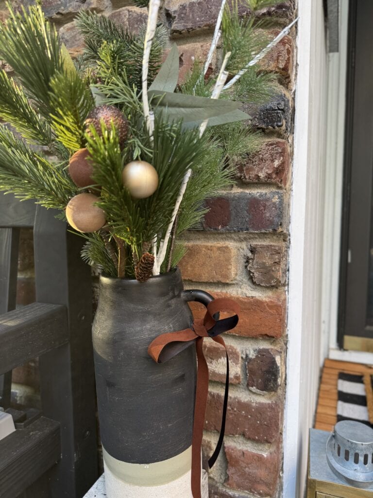 Festive holiday flower arrangement in black ceramic vase with ribbon, pine branches, ornaments, and greenery on porch with brick wall background.