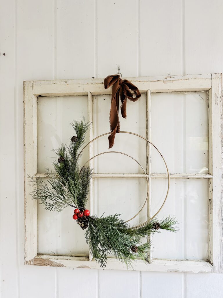 Rustic holiday wreath featuring pine branches, red berries, and pinecones, hung on vintage distressed window frame with brown cloth bow for Christmas decoration.