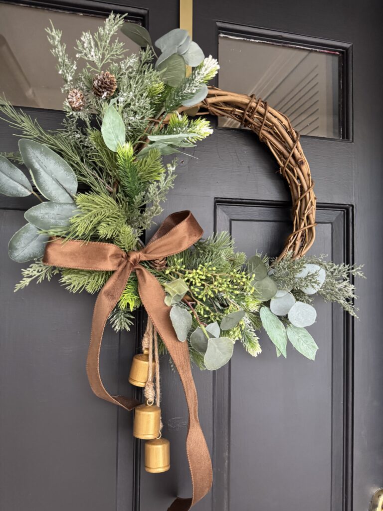 Lush holiday wreath with greenery, pinecones, eucalyptus, and a brown satin bow hanging on a black door for festive decor.