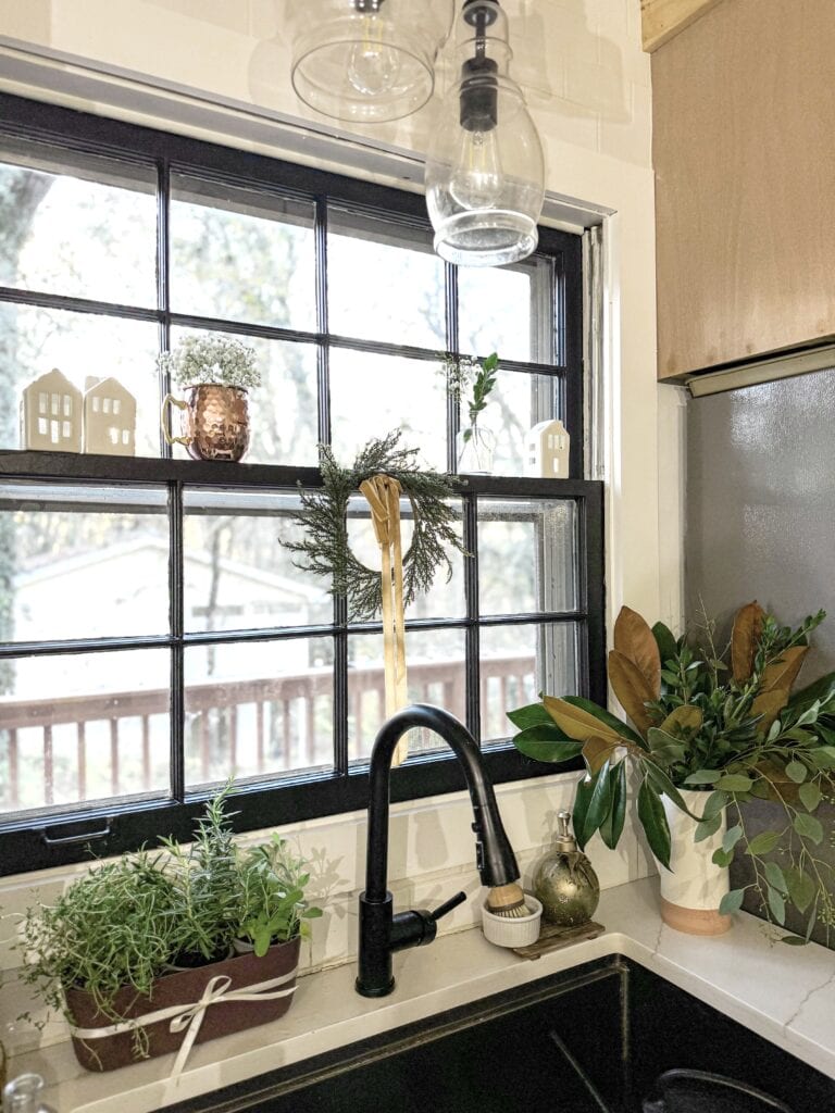 Cozy kitchen window with decorative plants and holiday wreath, black sink faucet, holiday decor, white countertop, and natural light, creating a warm and inviting kitchen ambiance.