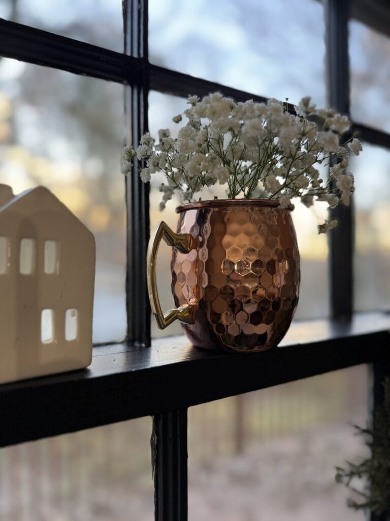 Shiny copper hammered mug with gold heart-shaped handle filled with white baby's breath flowers on a windowsill, cozy home decor.