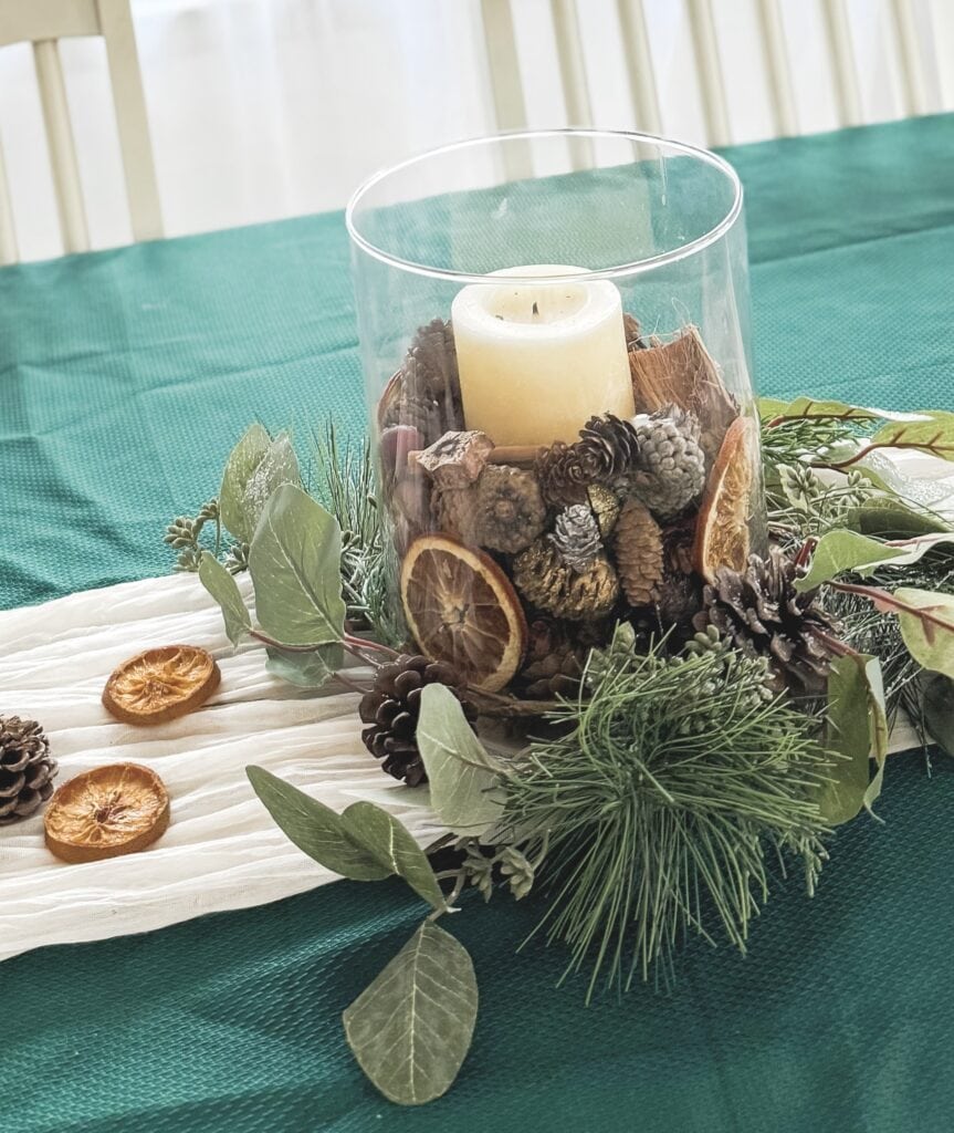 Rustic holiday centerpiece with pinecones, dried orange slices, and a large candle in a glass holder, perfect for winter decor and festive table settings.