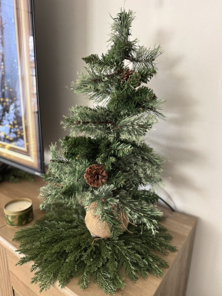 Lush miniature Christmas tree decorated with natural pine cones, placed on a wooden table with holiday greenery and a candle.