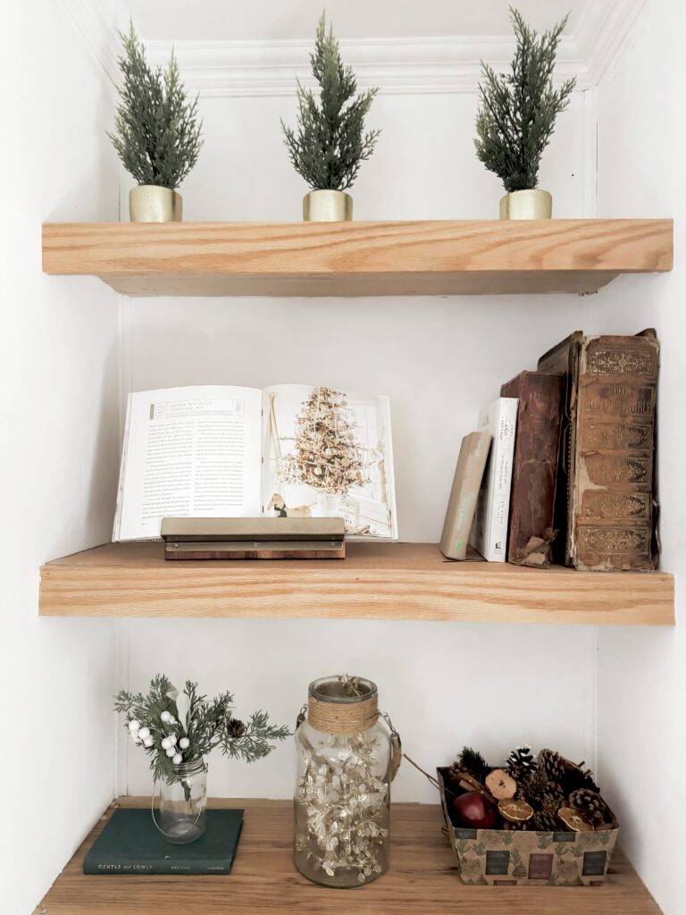 Mini Christmas trees in gold pots on upper wooden shelf, vintage books and an open holiday-themed book on middle shelf, decorative jar with fairy lights and festive ornaments on lower shelf.