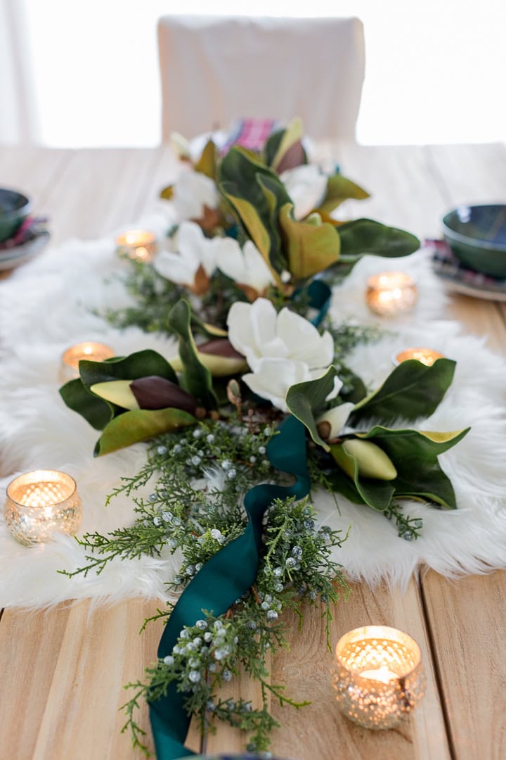 Elegant floral centerpiece with magnolia and greenery for special occasions, beautifully accented with teal ribbon, candles, and cozy table setting on a rustic wooden table.