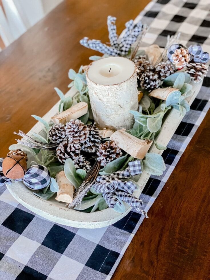 Birch candle centerpiece with pinecones, eucalyptus, and gingham ribbons on a rustic wooden table for cozy home decor or holiday table setting.