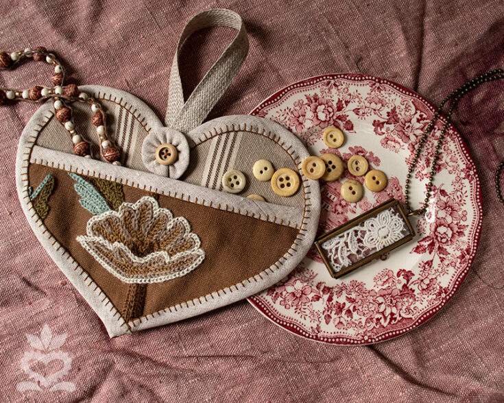 A heart-shaped embroidery kit featuring buttons, a floral embroidered design, and delicate jewelry displayed on vintage fabric, perfect for sewing and quilting enthusiasts.