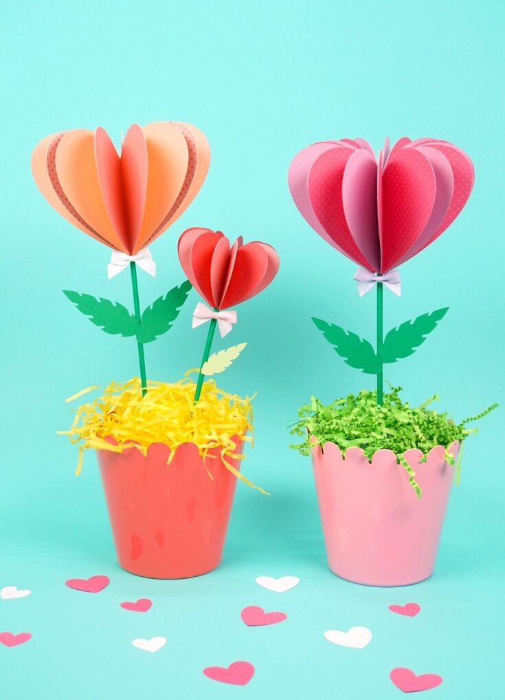 Bright pink and peach paper flowers in colorful pots with green leaves, paper grass, and heart-shaped confetti for love and affection gifting.