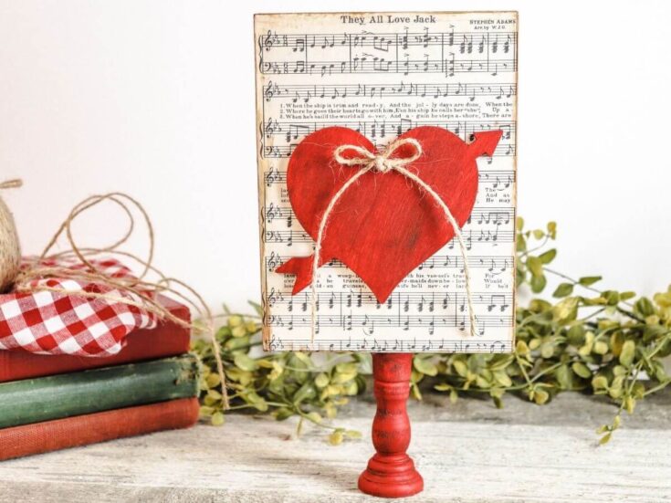 A charming red wooden heart attached to a vintage sheet music backdrop, perfect for romantic decor or wedding table decor, with greenery and stacked fabric gifts nearby.