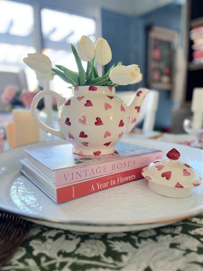 A charming white teapot decorated with pink and red hearts, filled with fresh white tulips, placed on top of vintage floral books, creating a cozy, romantic table setting at A Hundred Affections.