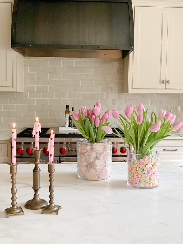 Beautiful pink tulips in glass vases filled with pastel candies and pink stones, paired with vintage candlesticks with lit candles, creating a charming and cozy kitchen display.