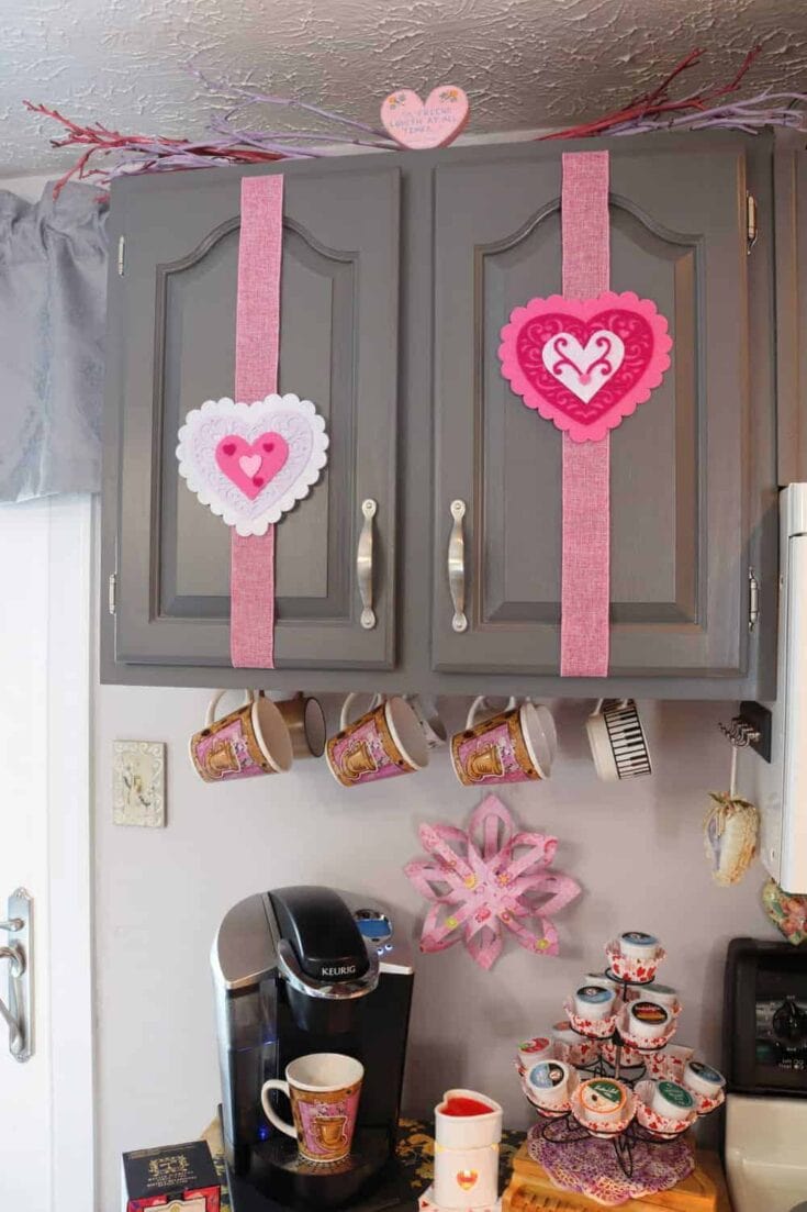 Decorative pink heart-themed Valentines Day kitchen wall hanging with coffee mugs and craft elements.
