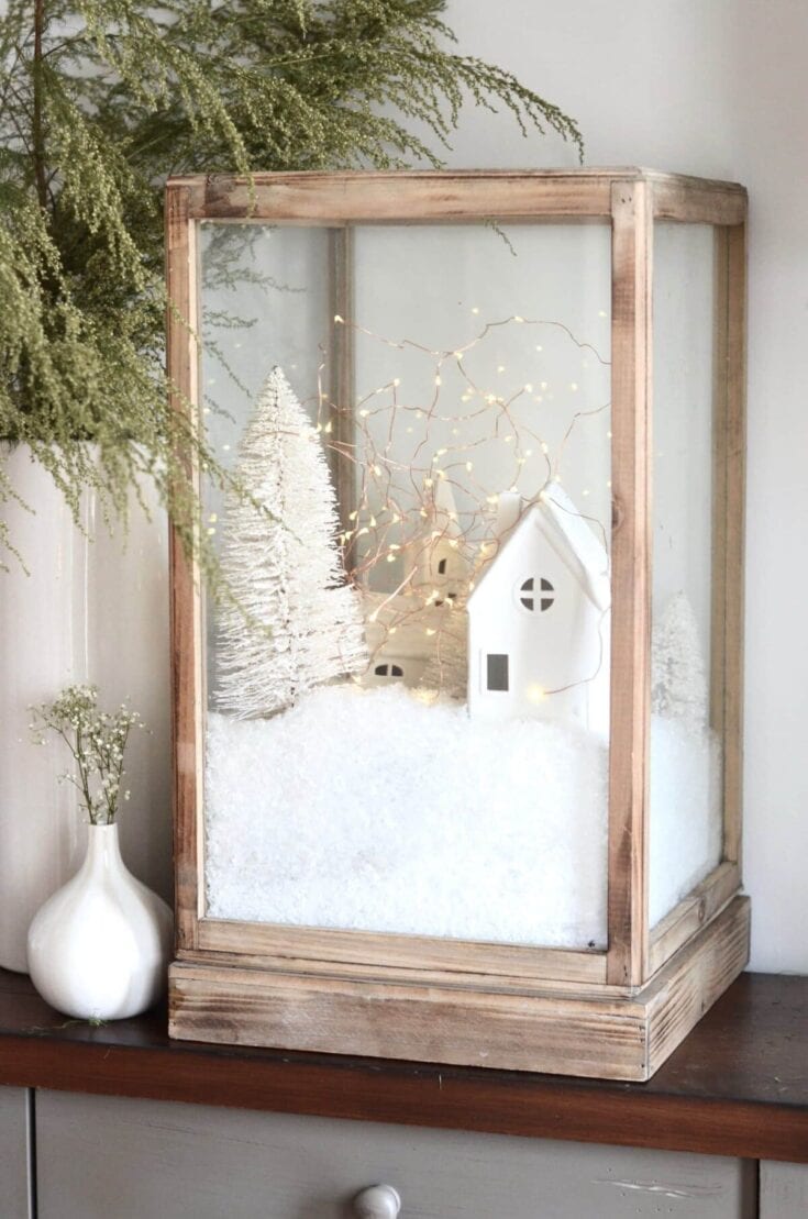 Elegant winter holiday display featuring a white ceramic house, snowy miniature trees, warm string lights, and festive greenery for cozy holiday decor.
