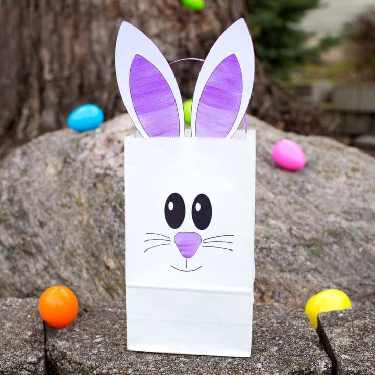 Cute Easter gift bag with bunny face and purple bunny ears, surrounded by colorful plastic eggs on rocky surface, perfect for festive celebrations and holiday gifting.