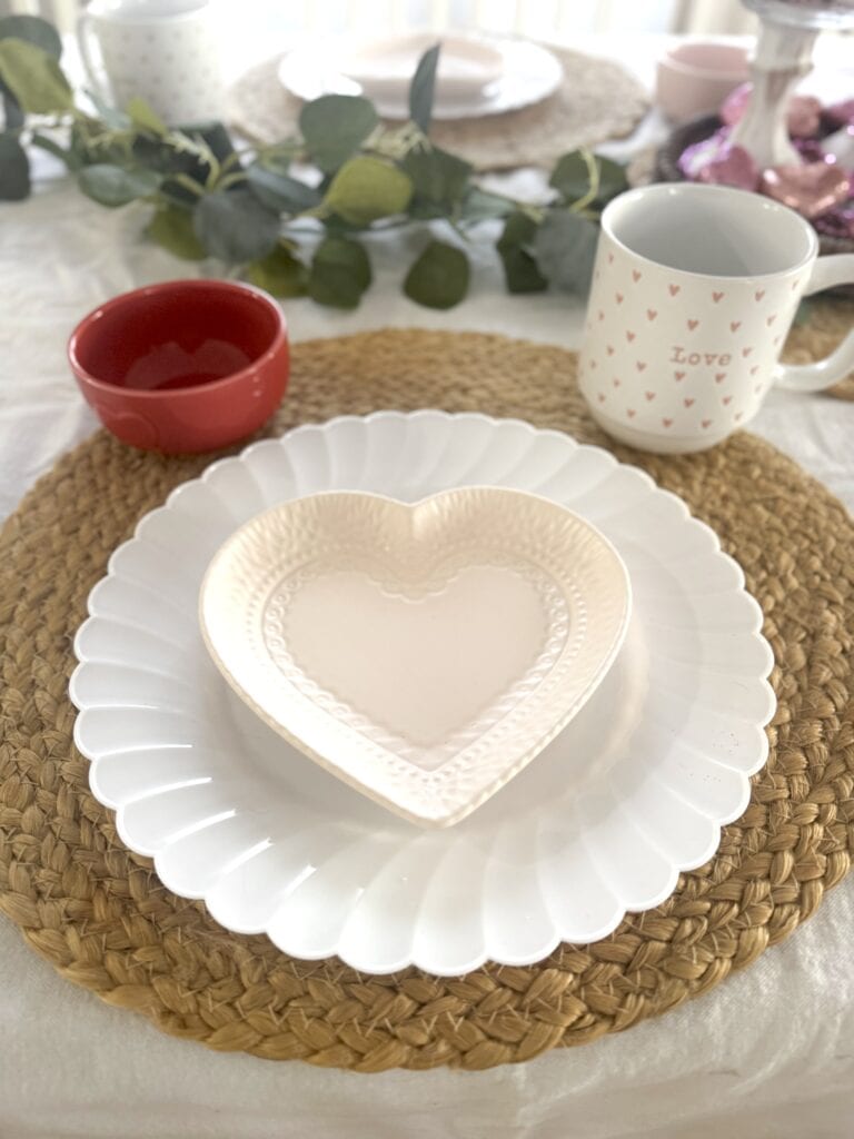 A charming breakfast table featuring a white scalloped plate with a smaller heart-shaped dish, surrounded by cheerful mugs, bowls, and greenery. Perfect for cozy gatherings and Valentine's Day.