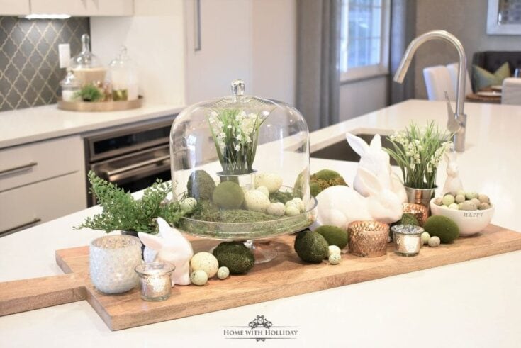Elegant Easter table centerpiece with ceramic bunnies, floral accents, moss, candles, and decorative eggs in a modern kitchen setting.