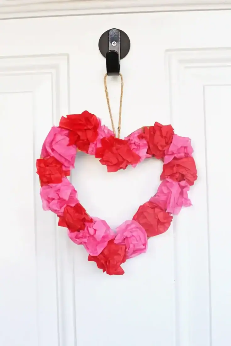 Colorful tissue paper heart wreath hanging on a white door, perfect for Valentine's Day or romantic decor. Bright pink and red tissue paper craft for love-themed home decoration.
