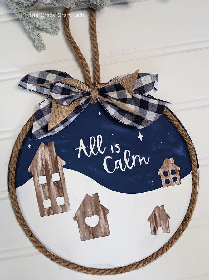 Handmade circular wall art with rustic houses and the phrase "All is Calm", featuring a gingham ribbon bow, perfect for cozy home decor and holiday seasonal decoration.
