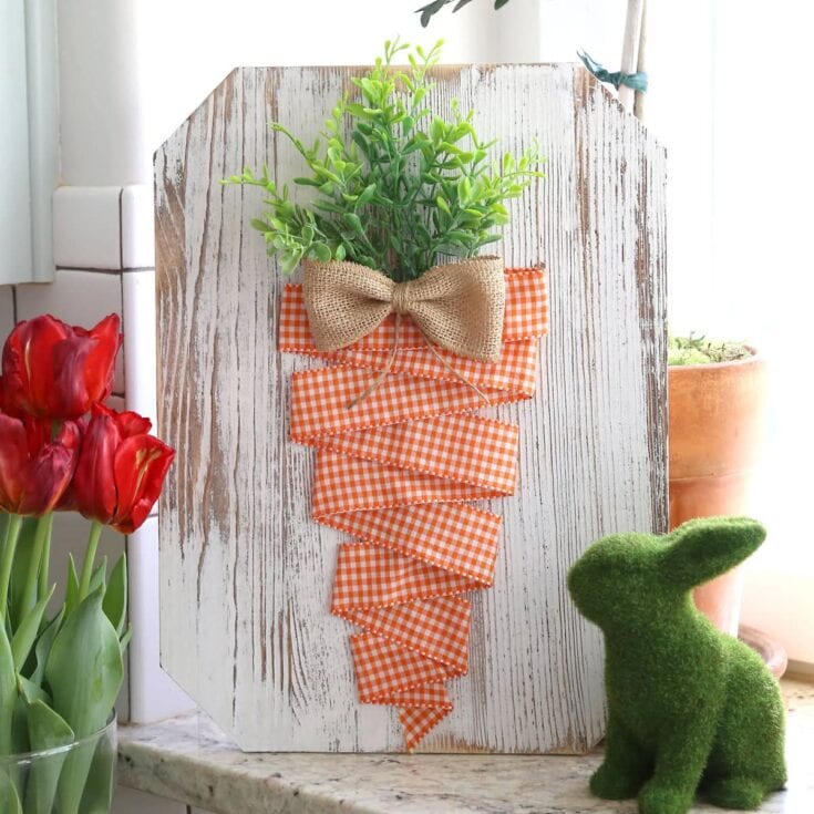 A decorative arrangement featuring a green plant wrapped in orange gingham fabric with a burlap bow, displayed on rustic white wood, perfect for spring or Easter celebrations.