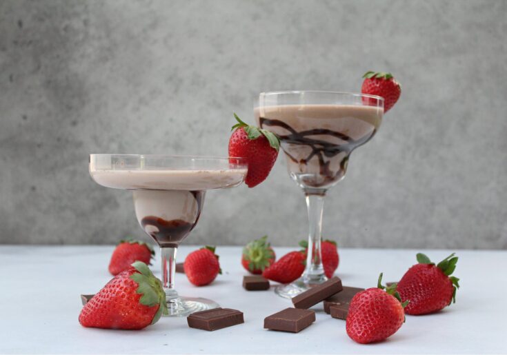 Elegant glass cups filled with creamy strawberry chocolate mousse garnished with fresh strawberries, surrounded by chocolate pieces and strawberries on a white surface, gray background.