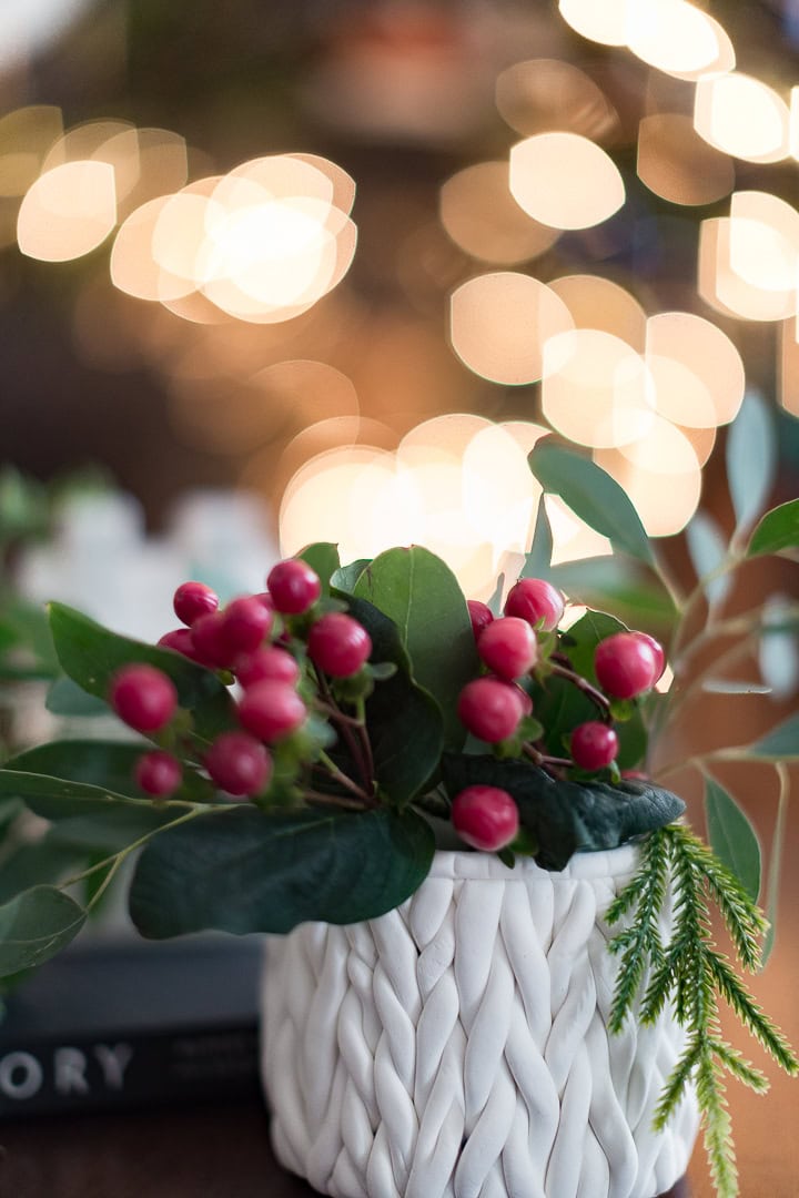 A festive arrangement featuring holly berries and green foliage in a textured white basket with blurred warm bokeh lights background, perfect for holiday celebrations and seasonal home decor.