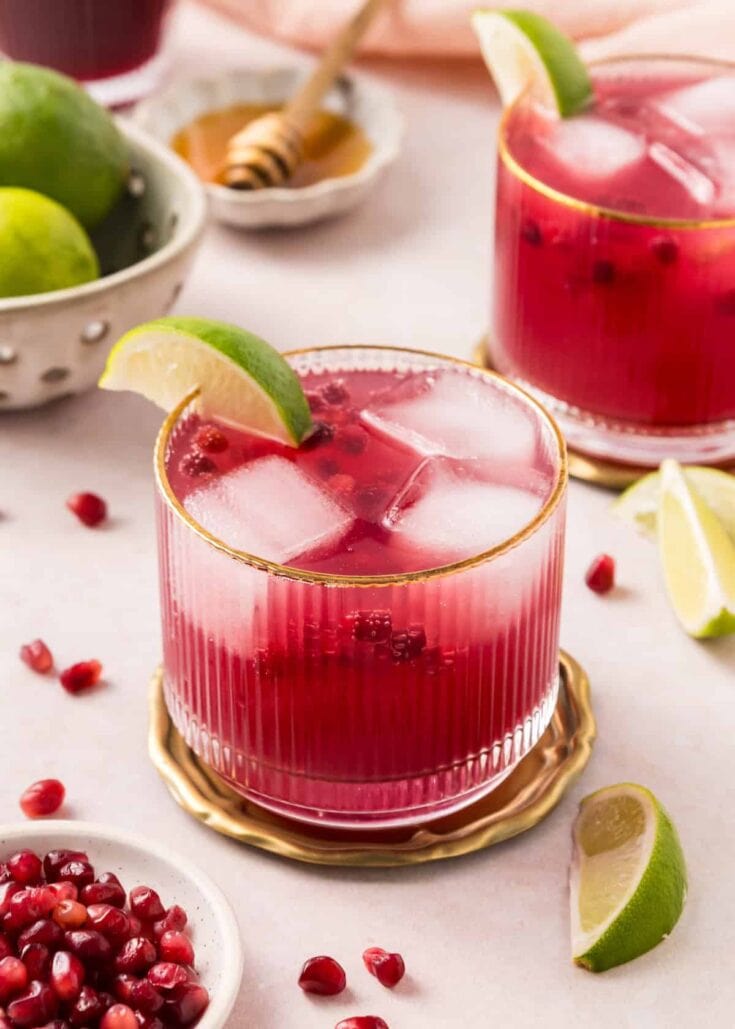 A vibrant glass of cranberry lemonade with ice, lime wedge garnish, and fresh pomegranate scattered around, perfect for summer refreshment and festive gatherings.