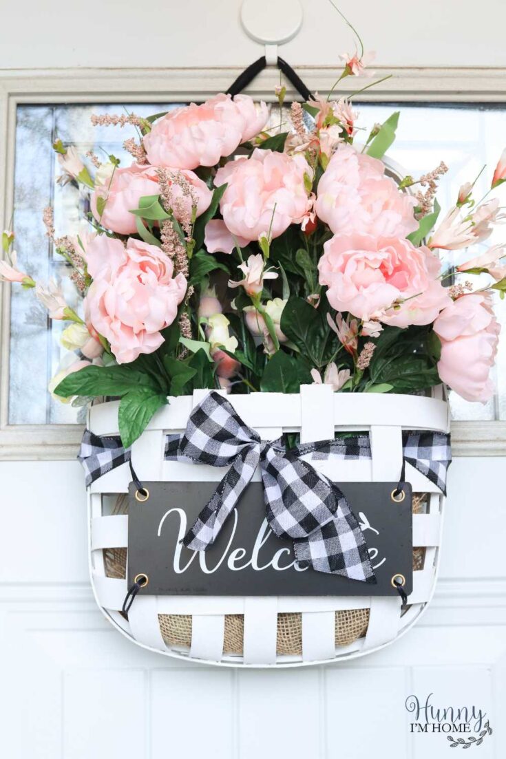 Beautiful pink peonies and greenery flower arrangement in a white wicker basket with "Welcome" sign and black-and-white checkered bow, perfect for home decor or gifting.