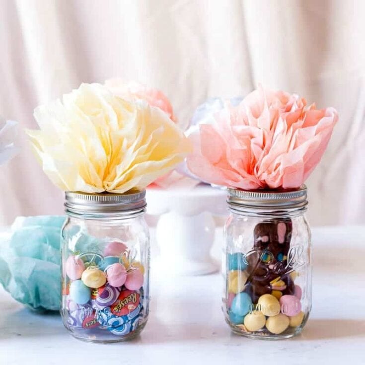 Colorful paper flowers in glass jars filled with assorted candies, creating a charming and whimsical decoration for celebrations or home decor.