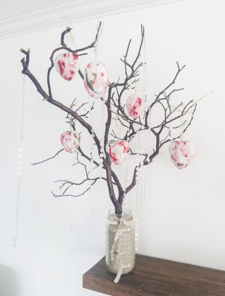 A decorative tree with heart-shaped ornaments painted in pink and white, displayed in a glass jar on a wooden surface, symbolizing love and affection.
