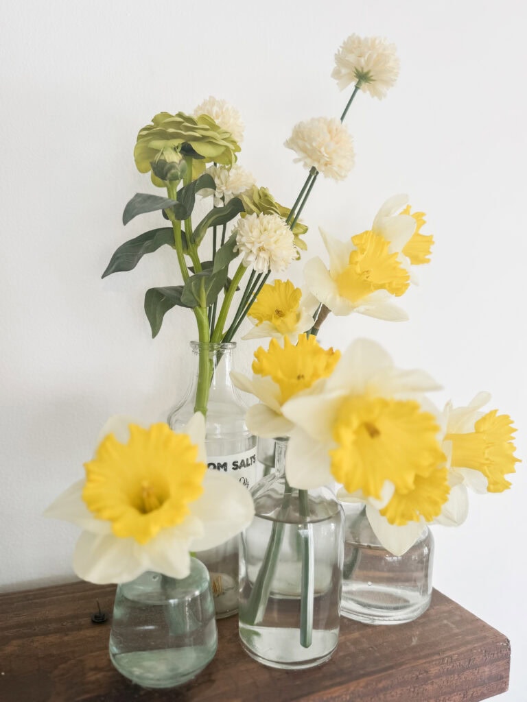 A collection of fresh yellow and white daffodils and white pom-pom flowers arranged in clear glass vases on a wooden surface, creating a bright and cheerful floral display.