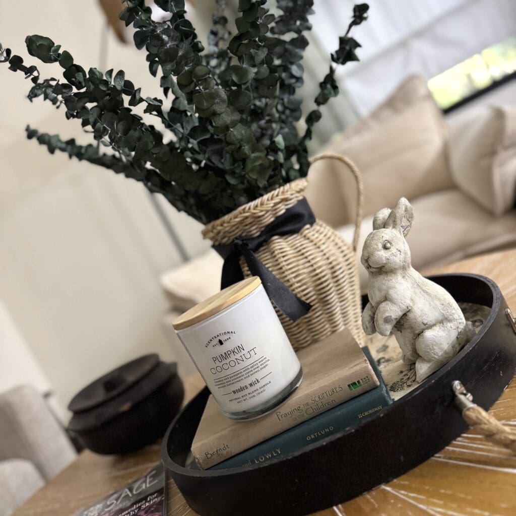 A cozy decorative arrangement featuring a pumpkin coconut scented candle, fresh eucalyptus, and a charming bunny figurine on a black tray, perfect for home decor.
