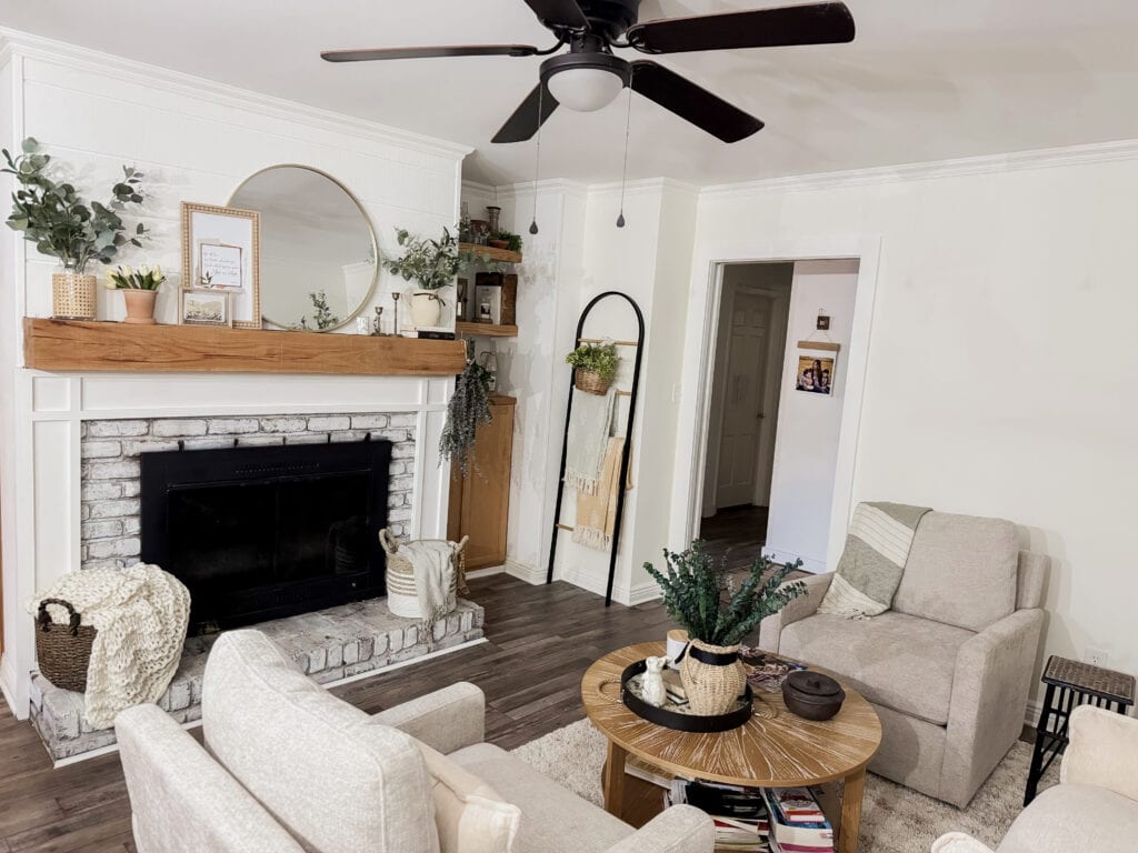 A welcoming living room featuring a white brick fireplace, wooden mantel with decorative items, neutral-toned sofas, and a round coffee table with plants and books, creating a warm, stylish space.