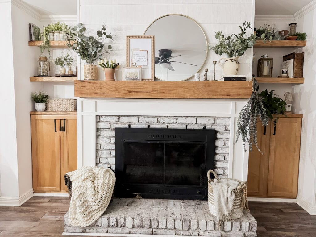 A cozy living room fireplace decorated with plants, framed art, and a round mirror, featuring wooden shelves and storage cabinets on either side for a warm, inviting atmosphere.