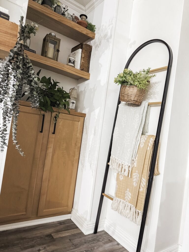 Stylish bathroom corner featuring a towel rack with decorative towels and a hanging plant, complemented by wooden shelves with plants and decor, creating a warm, inviting atmosphere.