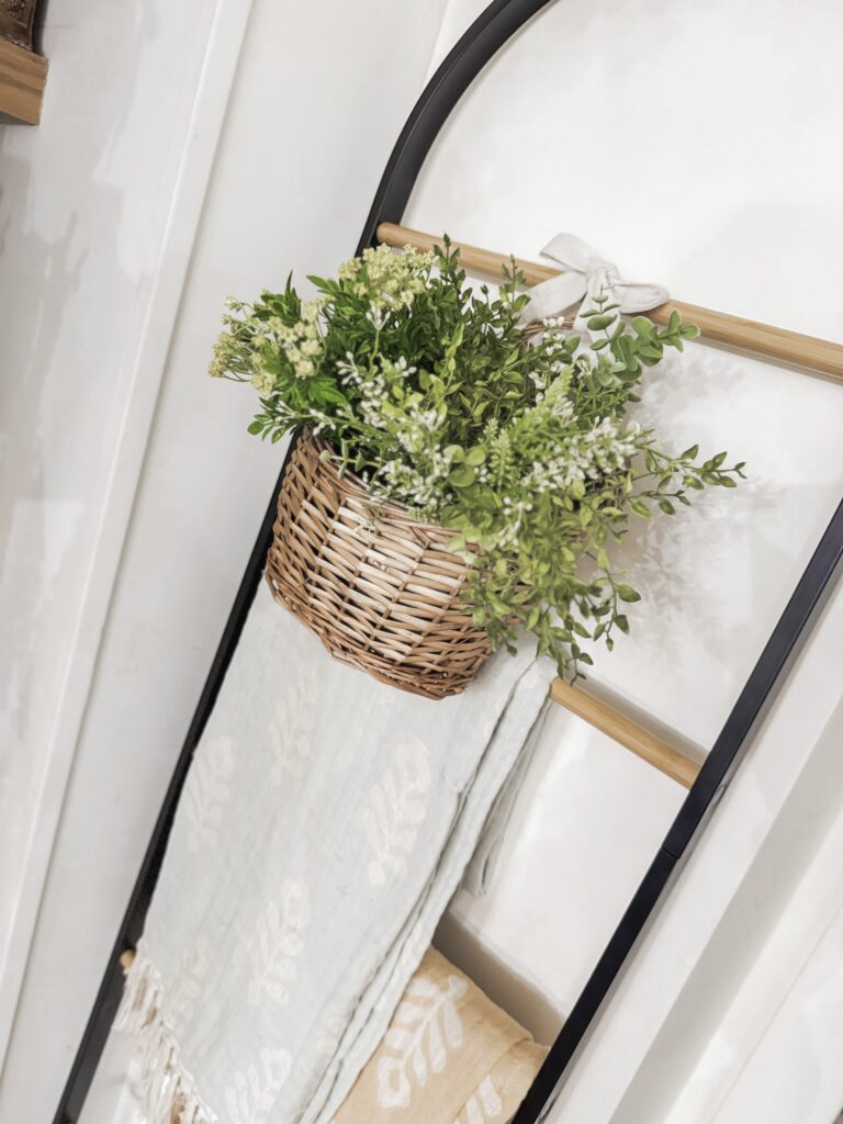 A decorative wicker basket filled with fresh green foliage and white flowers, hanging on a black metal stand in a cozy, well-lit interior space.