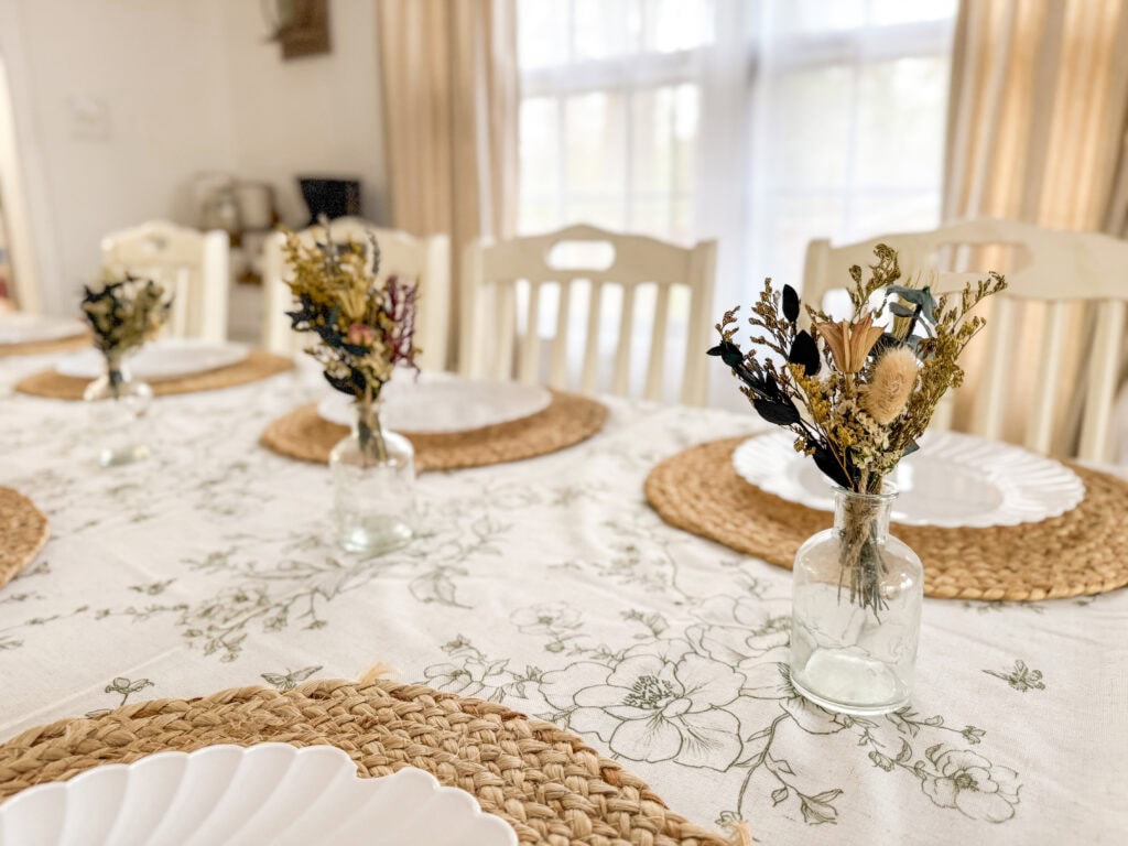 Elegant dining table setup with dried flower arrangements in glass jars, woven placemats, and a floral tablecloth, perfect for intimate gatherings and special occasions.