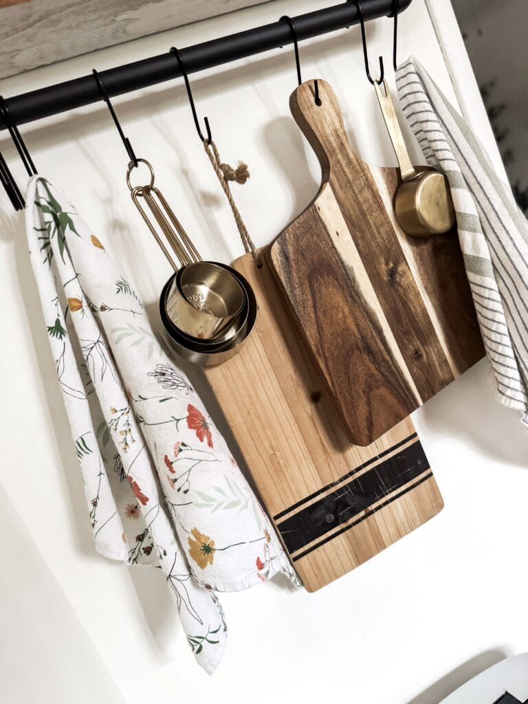 A set of wooden and bamboo chopping boards hanging on a kitchen rail with a brass measuring cup, a floral cloth, and a striped towel, ideal for kitchen organization and cooking prep.
