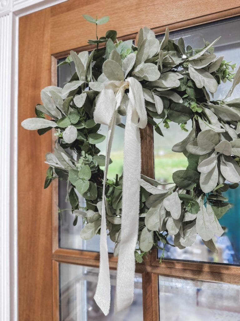 A beautiful greenery wreath made of lush, silvery-green leaves with a delicate fabric ribbon bow, perfect for home decor or seasonal decoration on a wooden door.