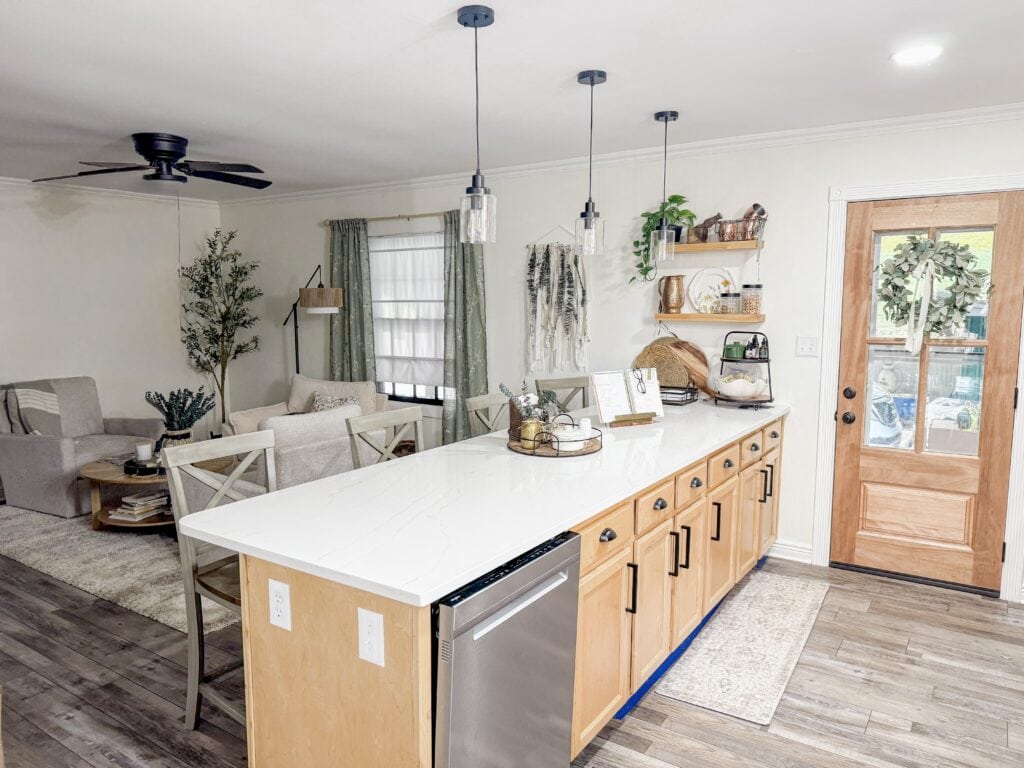 Bright and inviting kitchen featuring wooden cabinetry, open shelving with decorative items, and a spacious white countertop for cooking and gathering.