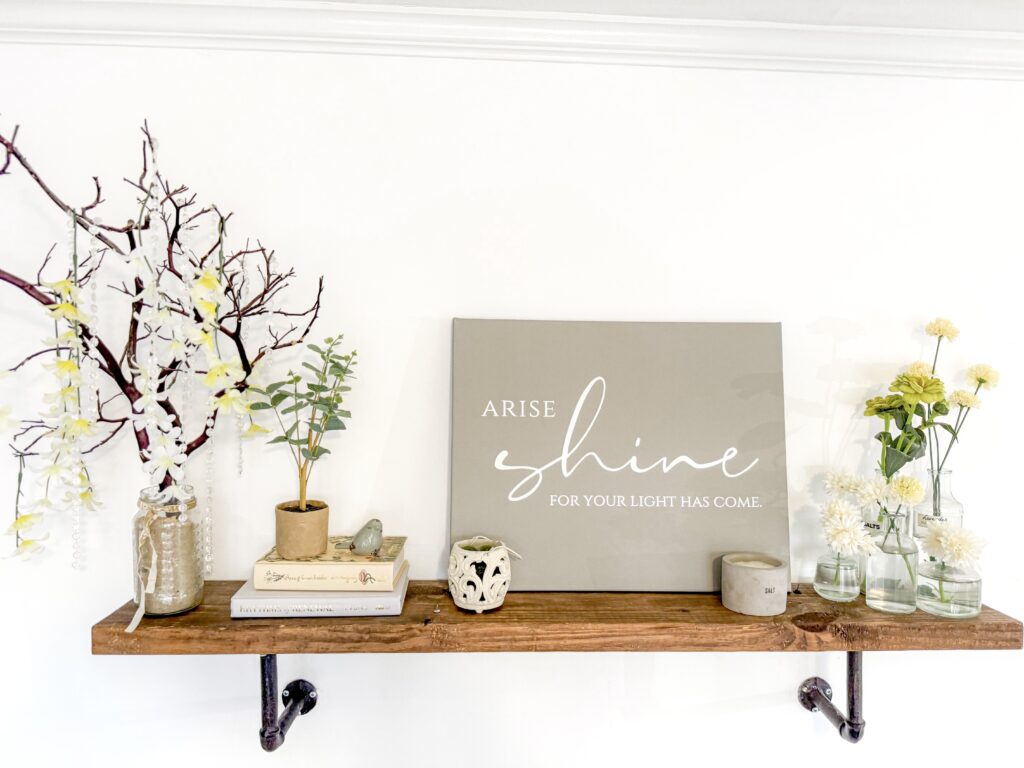 A decorative shelf with a neutral-toned "Arise Shine" sign, surrounded by vases with flowers, books, and candles, creating a cozy and inspiring home decor setup.