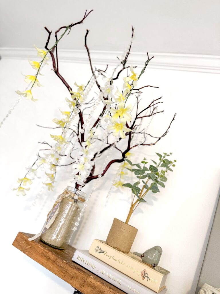 A decorative arrangement of yellow and white orchids with branches and green leaves in a rustic vase, placed on a wooden shelf for home decor and floral inspiration.