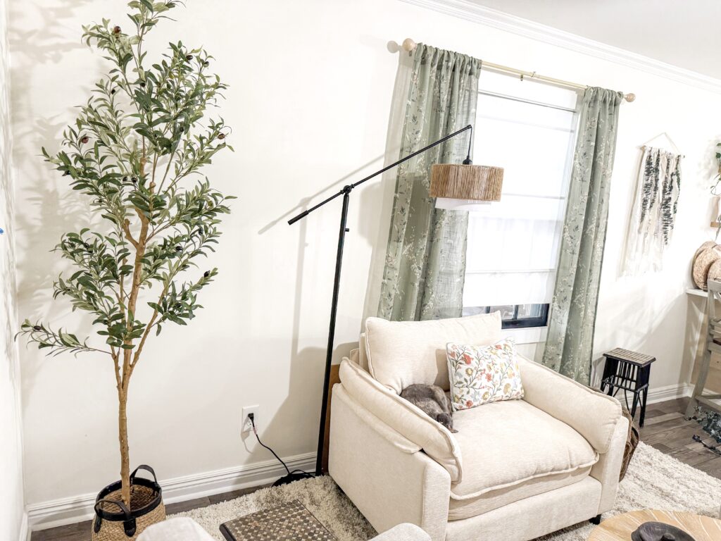 Bright and inviting living room featuring a beige sofa, large window with green curtains, indoor plant, and modern decor for a warm, relaxing atmosphere.