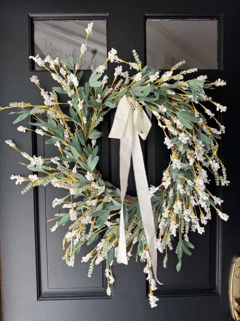 Beautiful spring floral wreath with white and yellow flowers, green leaves, and a white ribbon bow, hanging on a black front door, perfect for welcoming spring and adding charm to home decor.