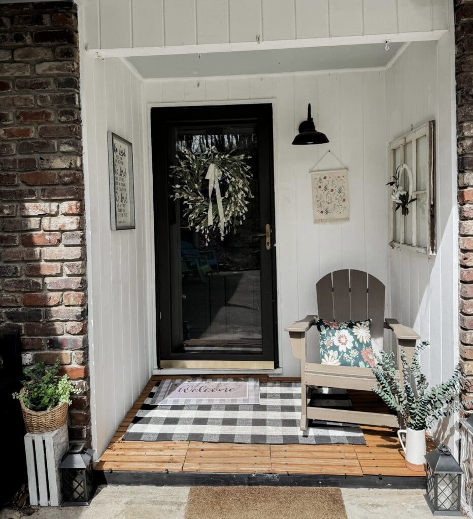 Front porch with white wood paneling, black door with wreath, Adirondack chair with floral cushion, potted plants, and rustic decor accents.