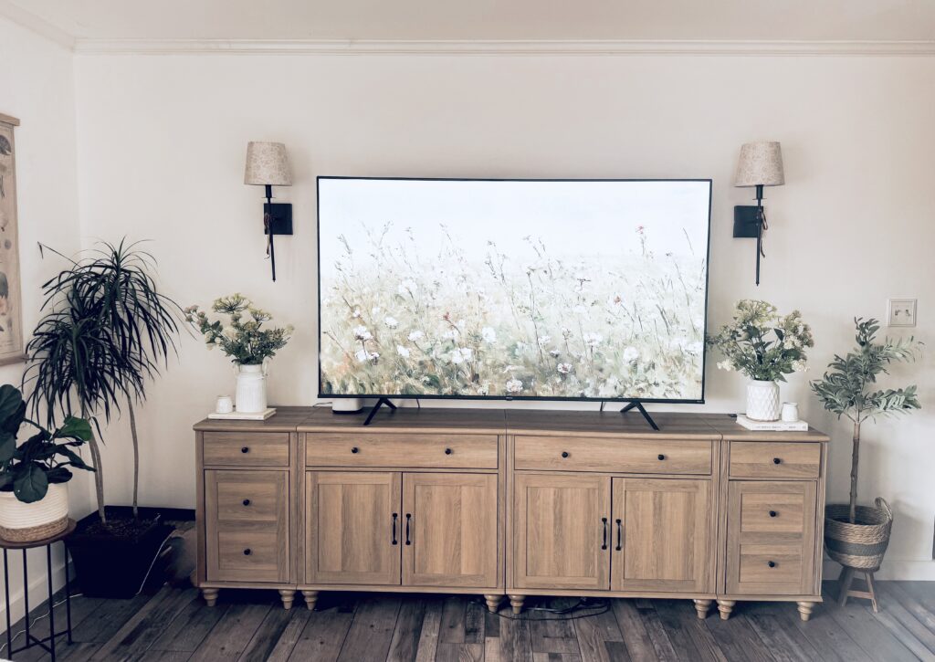 A cozy living room setup featuring a flat-screen TV on a wooden console, surrounded by potted plants and wall-mounted lamps, creating a warm and inviting atmosphere.
