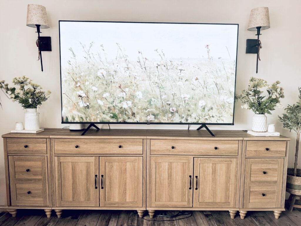 A large flat-screen TV displaying a nature scene with wildflowers, mounted above a wooden console table with potted plants and decorative lamps on either side, creating a cozy and natural ambiance.